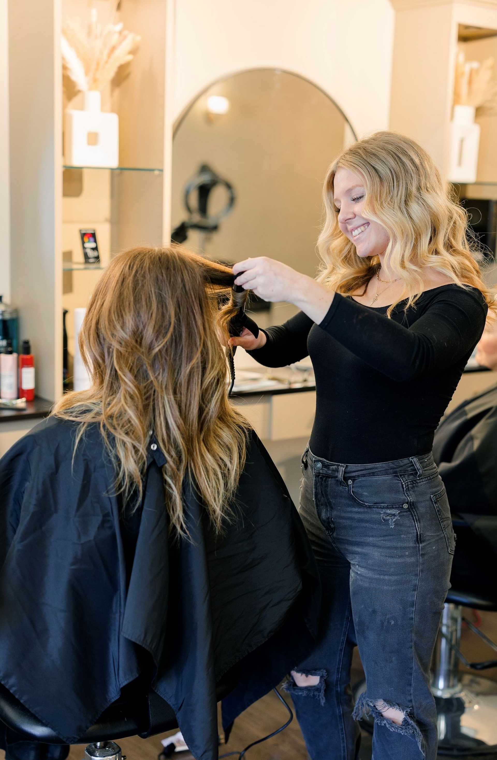 Stylist with long blonde hair smiling while curling client's hair in a modern salon.