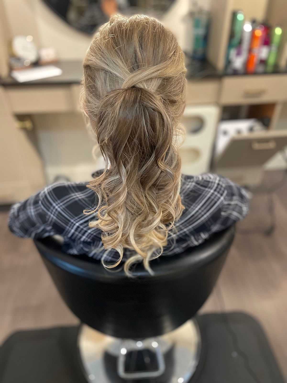 Blonde-haired woman with styled ponytail sits in a salon chair, facing away from the camera.